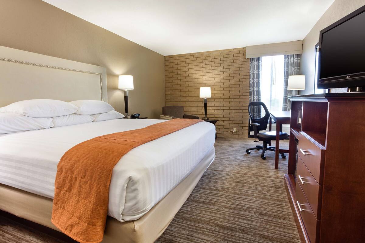A hotel room with a large bed, two bedside lamps, a desk with a chair, a television, and a brick-patterned wall.