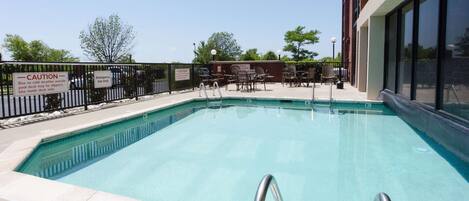 A swimming pool with a safety sign, poolside chairs, and a building in the background.