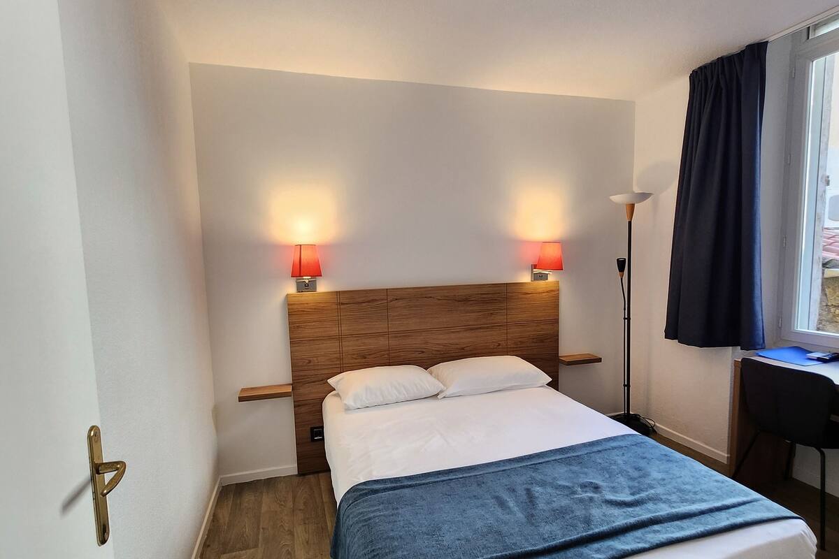 A hotel room with a wooden headboard, a bed with white and blue bedding, two bedside lamps, a desk with a chair, and a window with curtains.