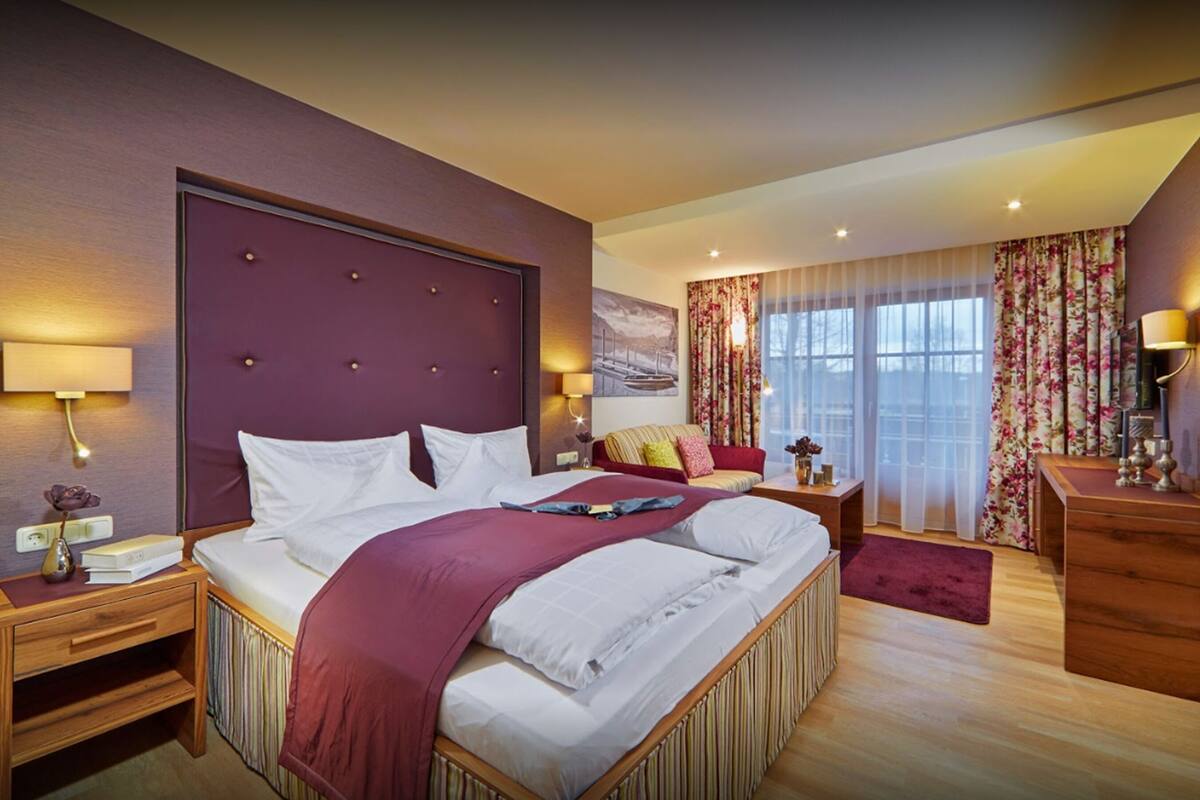 A hotel room with a large bed, a wooden nightstand, a desk with a lamp, and a window with curtains.