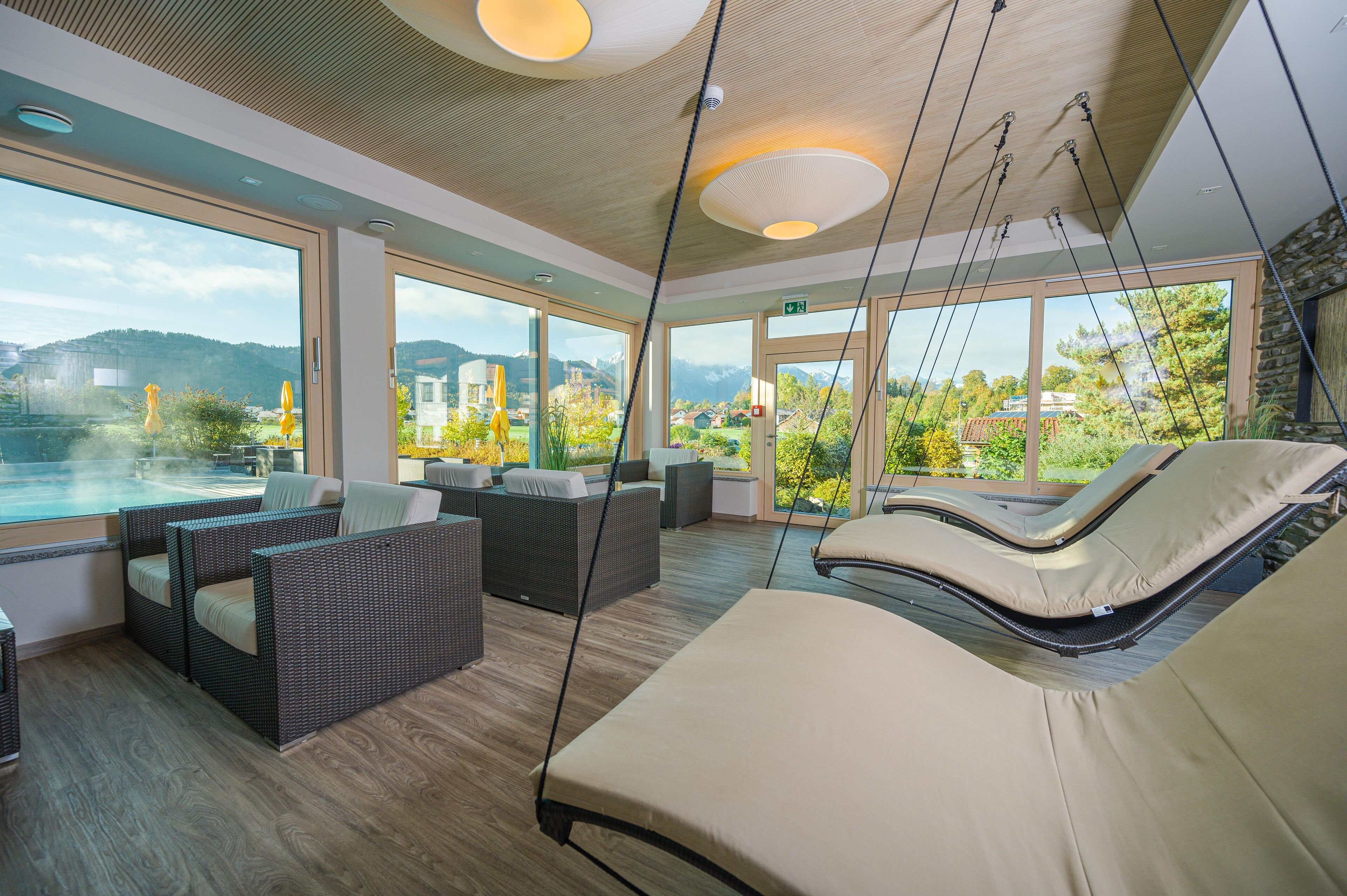 Un área interior moderna para relajarse, con muebles de mimbre, una sillona colgante y ventanas amplias con vistas a un paisaje montañoso.