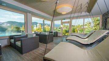 Un área interior moderna para relajarse, con muebles de mimbre, una sillona colgante y ventanas amplias con vistas a un paisaje montañoso.