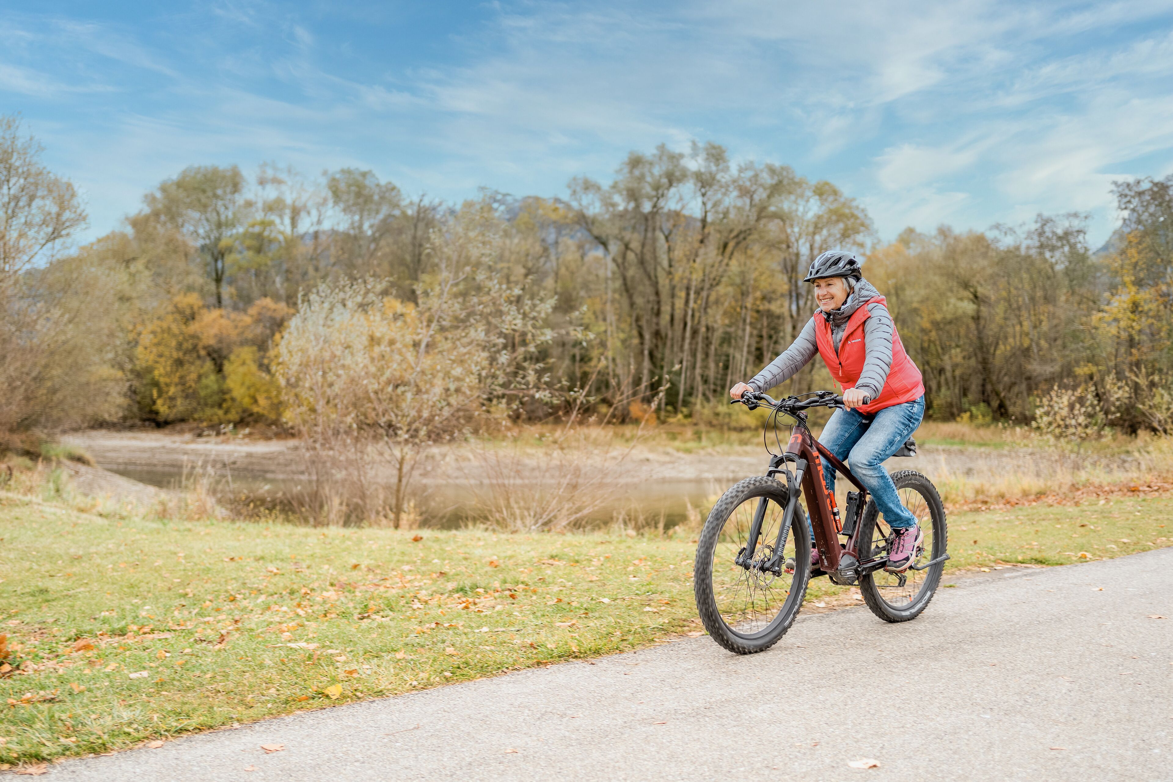 Una persona monta en bicicleta de montaña por un camino pavimentado en un parque con árboles y un cuerpo de agua al fondo.