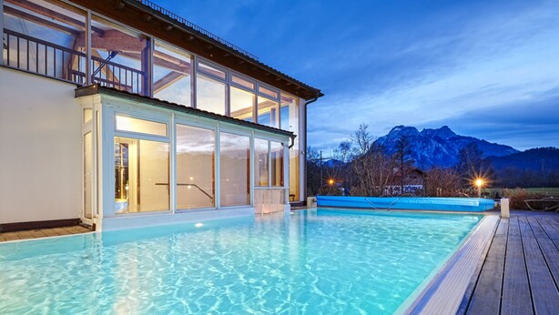 A swimming pool with a wooden deck, a glass-enclosed building, and mountains in the background. - Hotel Sommer Füssen (Fuessen)