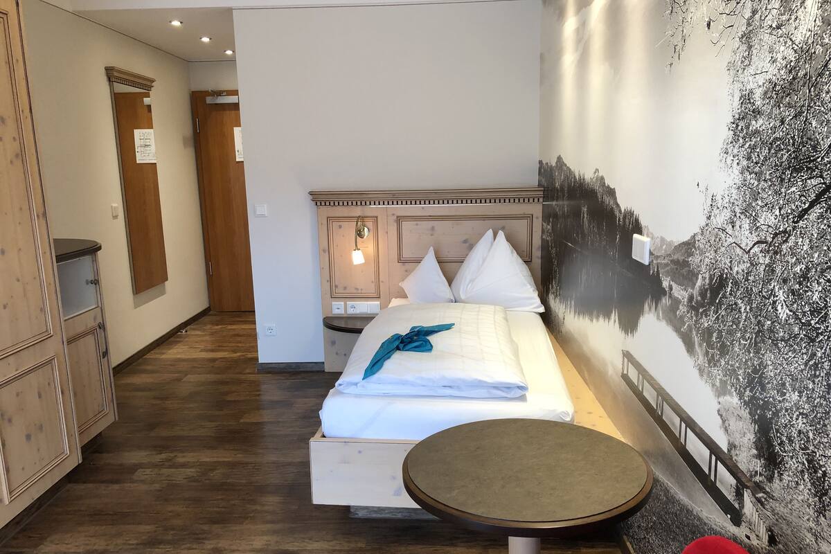 A hotel room with a bed, a small table, a wardrobe, and a wall with a mountain landscape mural.