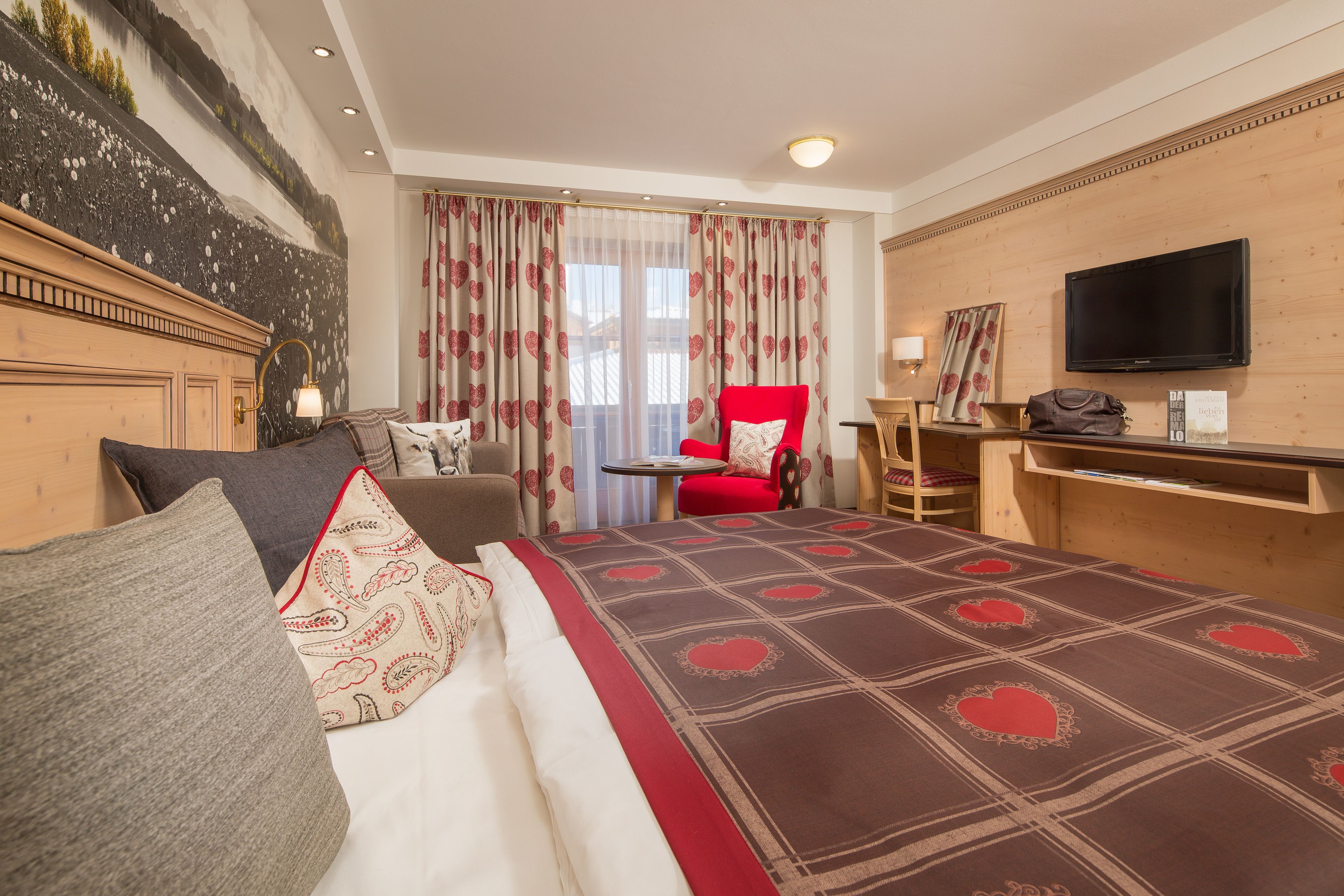 A hotel room with a bed, a red armchair, a TV, and a desk with a lamp.