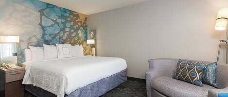 Una habitación de hotel con una cama, una silla, una mesita de noche, una lámpara y un cuadro en la pared.