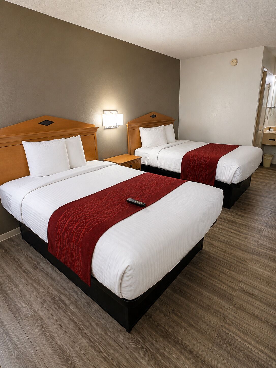 Une chambre d’hôtel avec deux lits, chacun doté d’une tête de lit en bois, de draps blancs et d’un plaid rouge. Il y a une table de chevet avec une lampe entre les deux lits, et le sol est en bois.