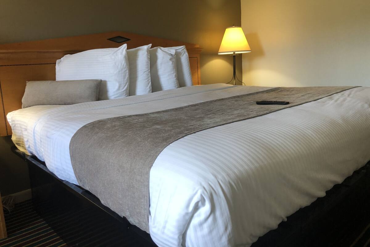 A neatly made bed with white linens and a grey throw, a bedside lamp, and a remote control on the bed.