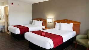 Quarto de hotel com duas camas, uma mesa de cabeceira, uma cadeira e uma luminária na parede.