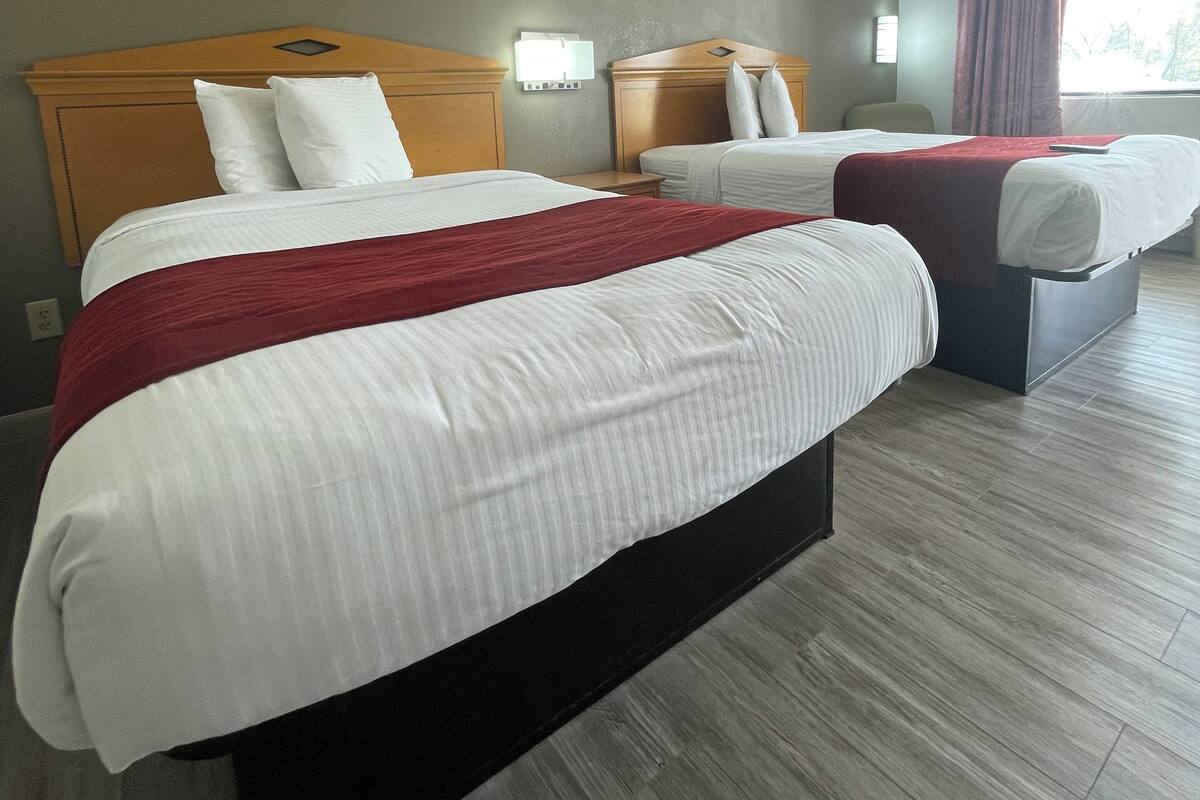 A hotel room with two beds, a wooden headboard, a red throw blanket, and a window with curtains.