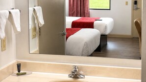 Quarto de hotel com um espelho grande, pia, duas camas, janela com vista para áreas verdes e porta com tranca.