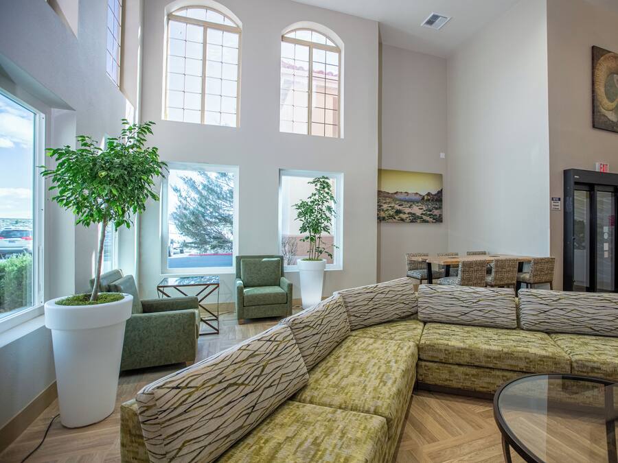 A spacious lobby with a large sectional sofa, two potted plants, a dining area with wicker chairs, and a painting on the wall.