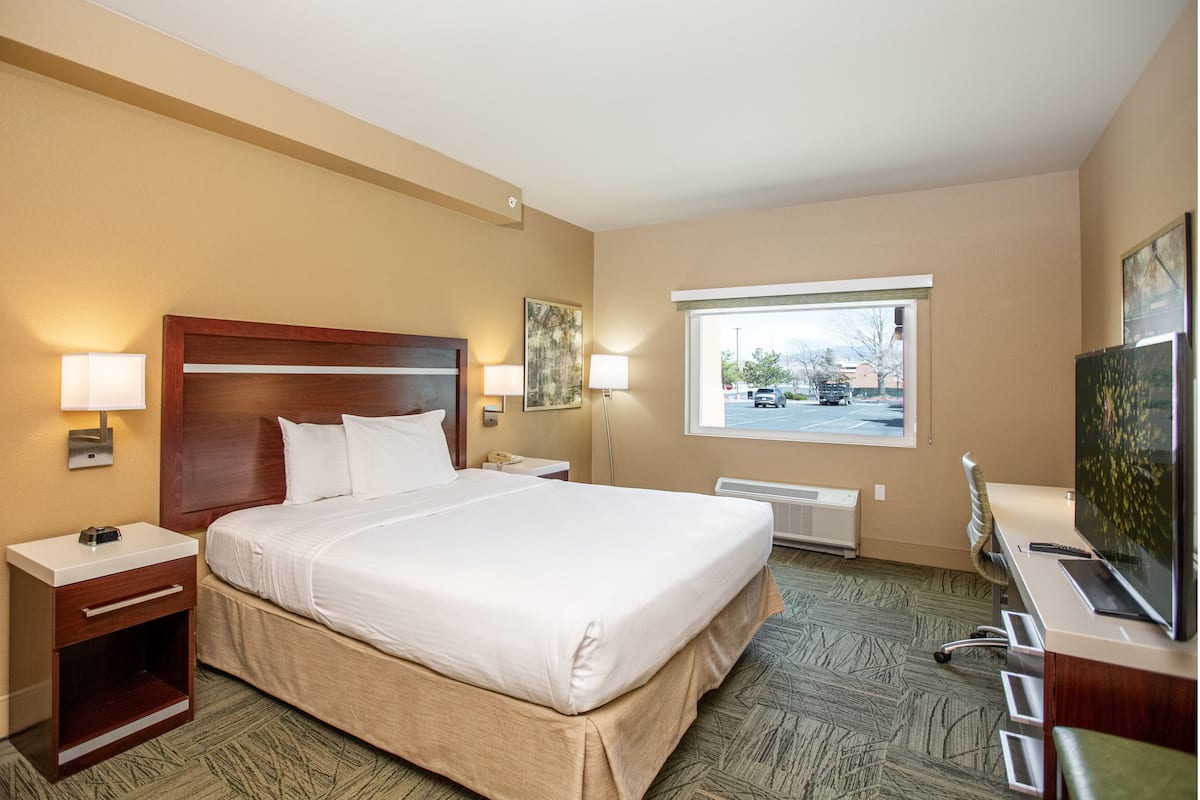 A hotel room with a large bed, two bedside tables with lamps, a desk with a computer and a TV, and a window with a view of the parking lot.