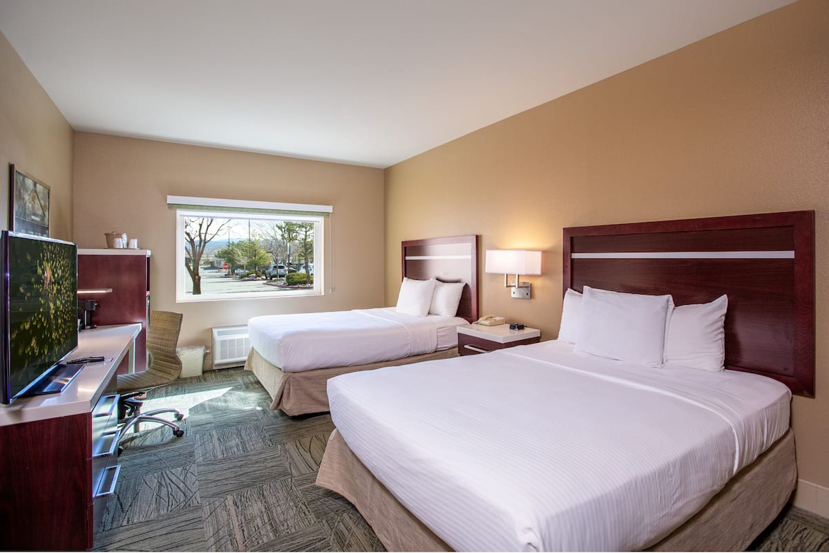 A hotel room with two beds, a desk with a TV, a window with a view of trees, and a lamp on the nightstand.
