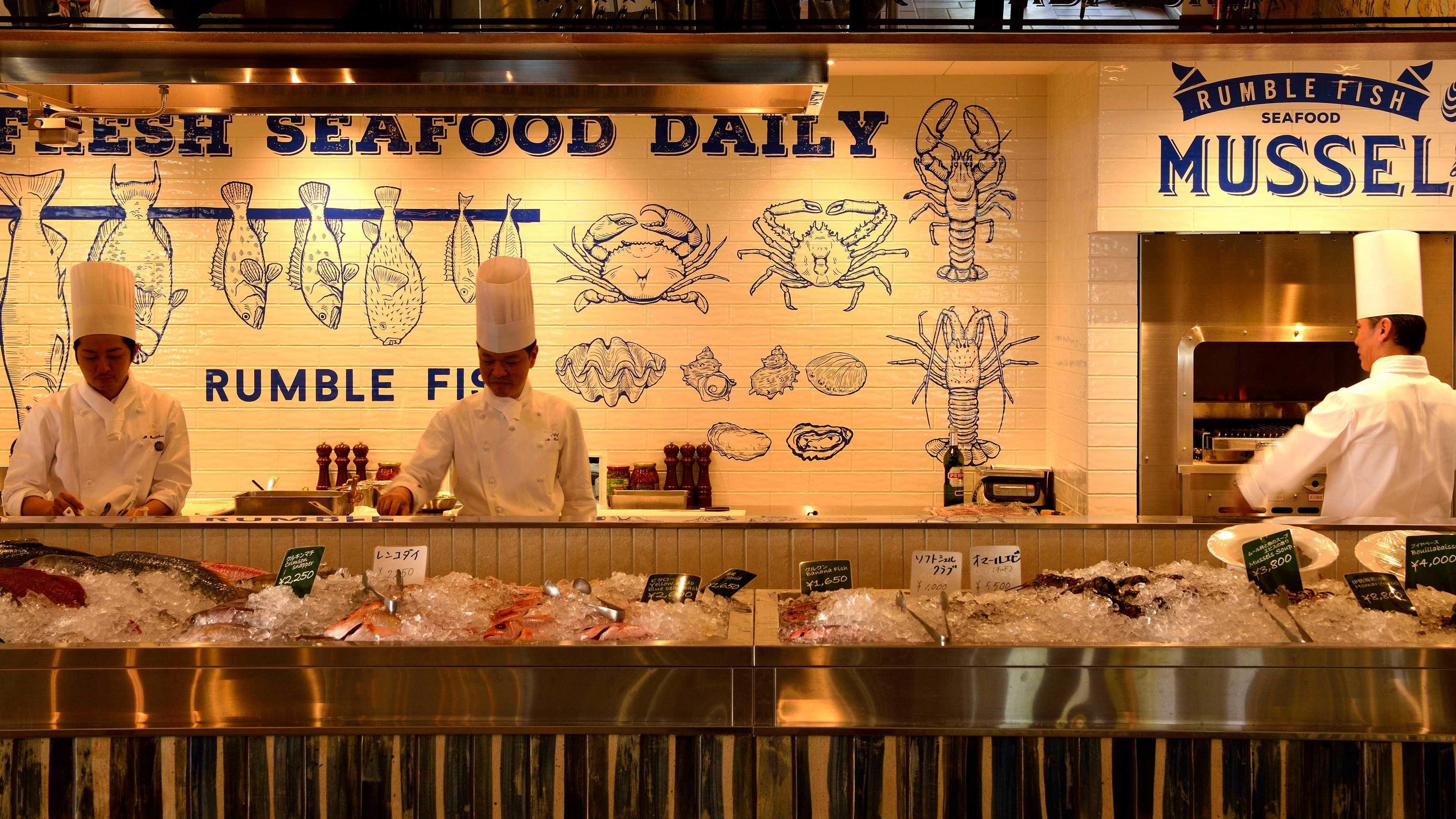 Mostrador de mariscos con chefs preparando productos frescos del mar, como mejillones, y una pared decorada con ilustraciones de peces.