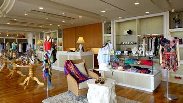 A boutique hotel interior with a central reception desk, display of clothing and accessories, and a seating area with a wicker chair and a small table.