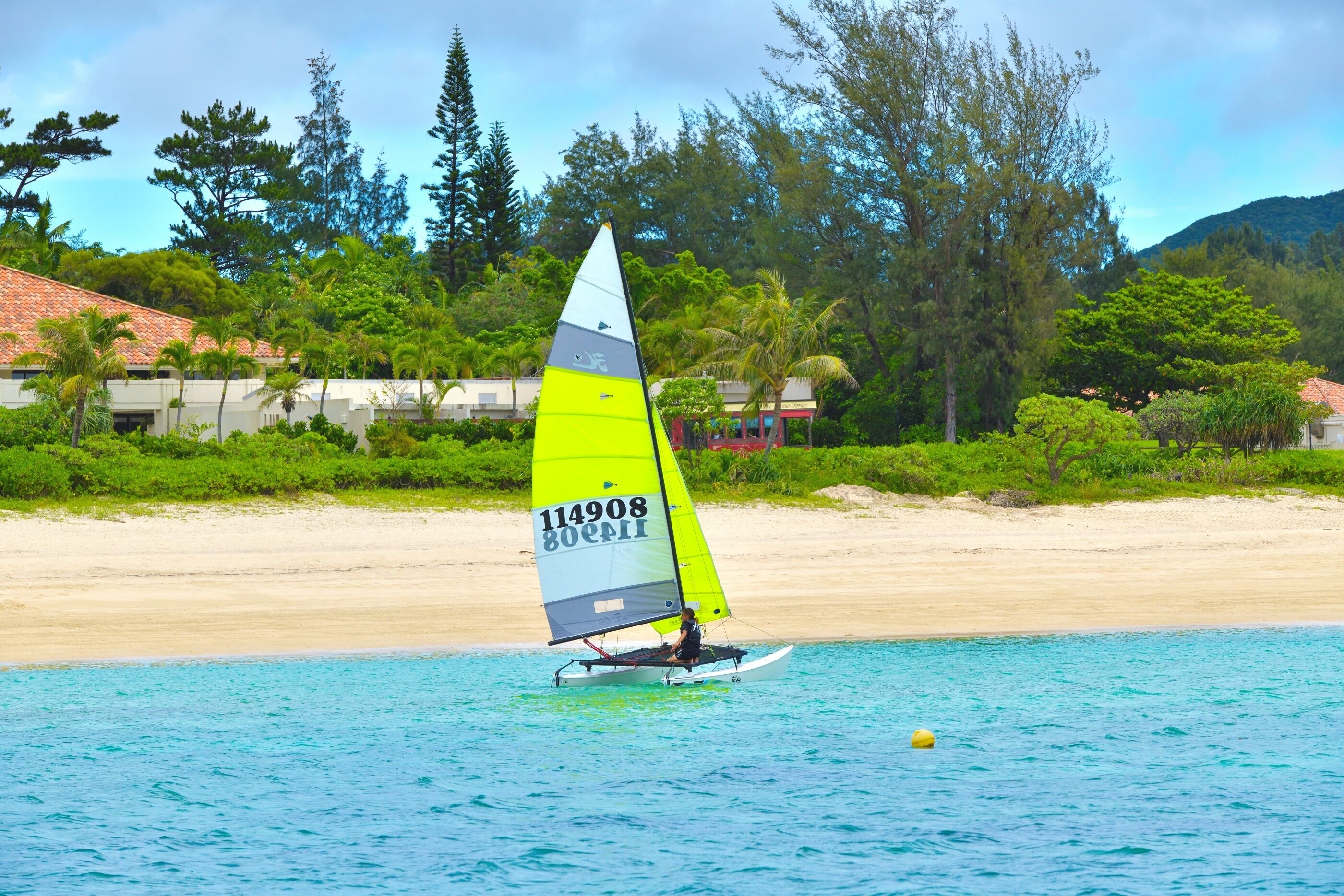 A sailboat with a yellow sail on calm water near a beach with houses and trees.