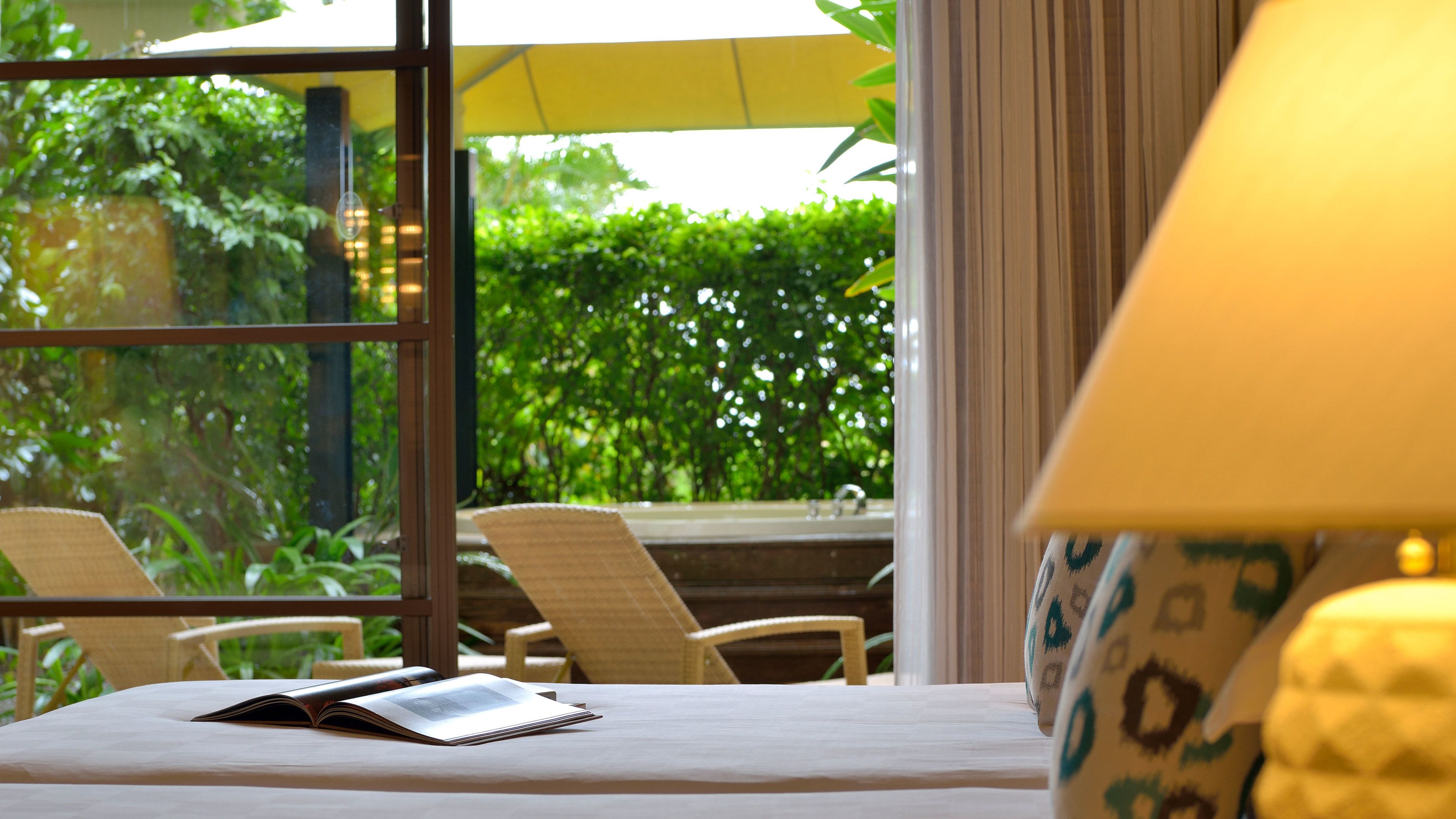 A hotel room with a bed, a window with a view of greenery, and a table with a book and a lamp.