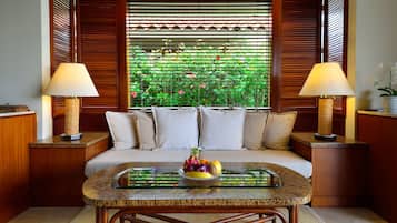 A room with a sofa, two lamps, a glass-top table with fruit, and a view of greenery through wooden blinds.