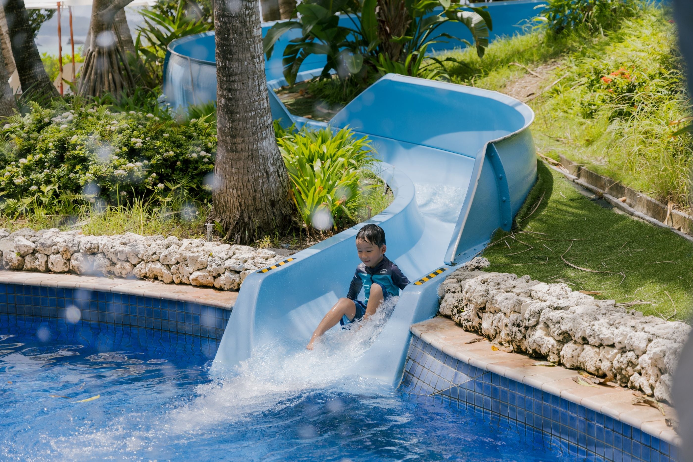 A child sliding down a blue water slide into a swimming pool surrounded by greenery.