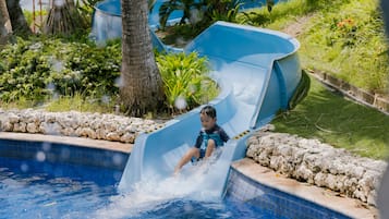 A child sliding down a blue water slide into a swimming pool surrounded by greenery.