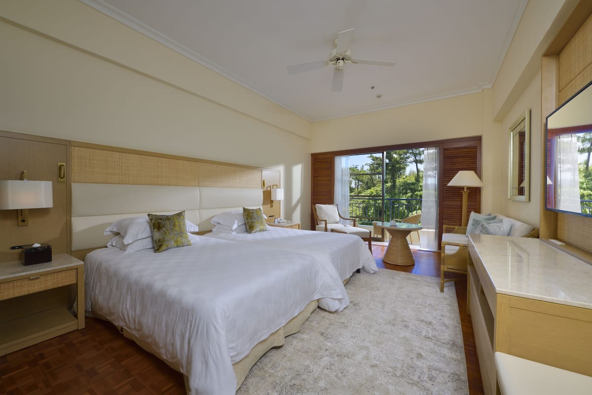 Ein Hotelzimmer mit zwei Betten, einem Schreibtisch, einem Stuhl, einem kleinen Tisch und einem Balkon mit Blick auf Grünflächen.