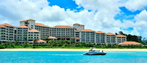 A large resort hotel with multiple buildings, a beach in the foreground, and a boat on the water.
