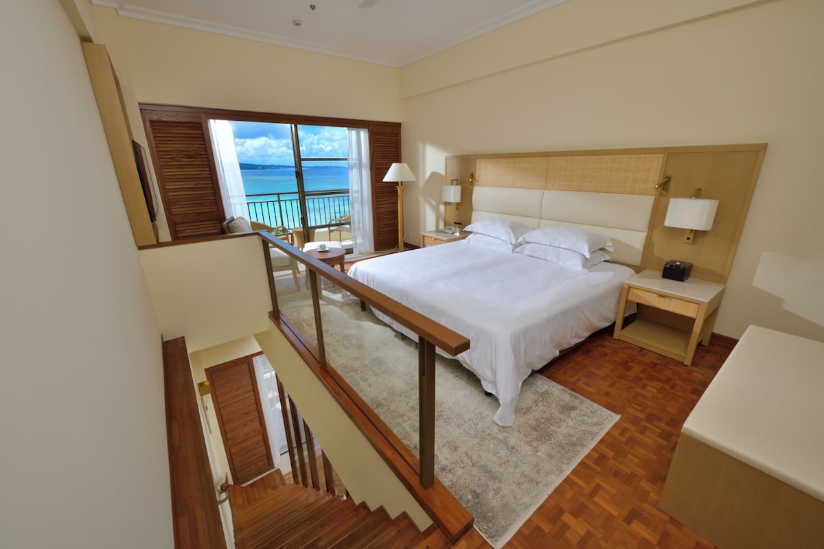 Ein Hotelzimmer mit einem großen Bett, einem Nachttisch, einem Schreibtisch und Meerblick durch eine Balkontür.