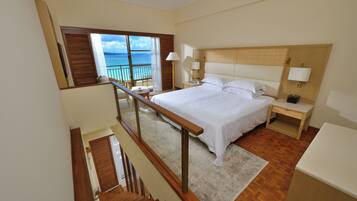 Habitación de hotel con una cama grande, una mesita de noche, un escritorio y vista al océano a través de una puerta de balcón.