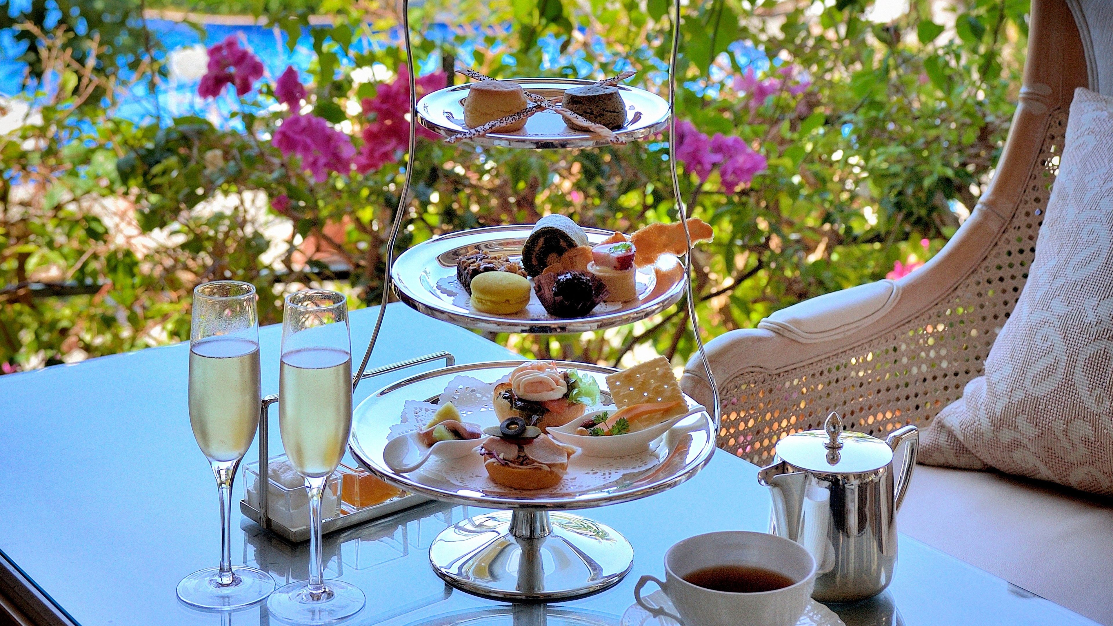 A tiered dessert tray with various sweets and pastries, two champagne flutes, a cup of coffee, and a silver teapot on a table with a view of greenery and flowers.