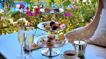 A tiered dessert tray with various sweets and pastries, two champagne flutes, a cup of coffee, and a silver teapot on a table with a view of greenery and flowers.
