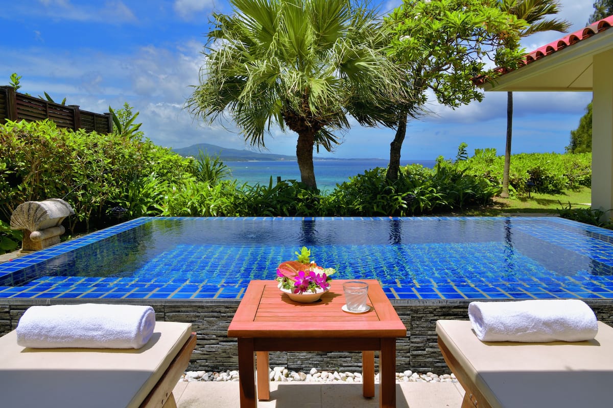 Ein Pool mit freiem Blick auf das Meer, umgeben von üppiger Vegetation und einem Holztisch mit Blumenarrangement.