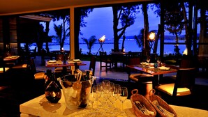 Un comedor con mesas dispuestas para una comida, un amplio ventanal con vista a la playa y la vista del océano al atardecer.