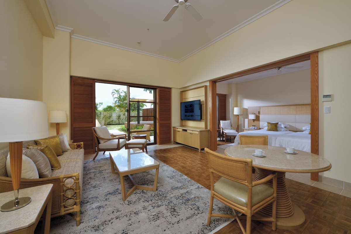 Ein Hotelzimmer mit Sofa, Stühlen, Esstisch, Fernseher, Bett und Blick ins Freie.