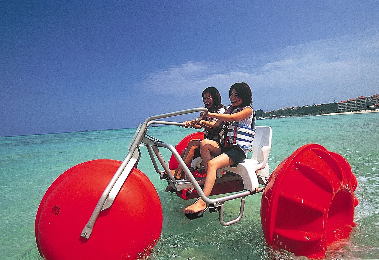 Two people on a red watercraft with a clear view of the ocean and a distant shoreline.