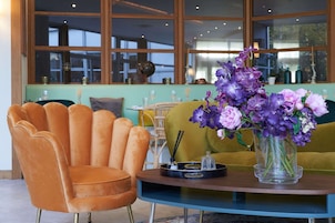 Una habitación con un ventanal grande, una mesa con un jarrón de flores, un sofá amarillo y un sillón naranja antiguo.
