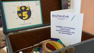 A suitcase containing a card with the text "BALTHAZAR ACADEMY" and "ANIMER LES BONNES PRATIQUES PROFESSIONNELLES AU QUOTIDIEN" along with some stationery items.