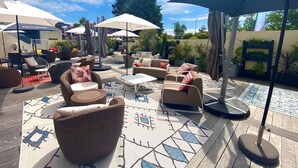 Un espace extérieur aménagé en terrasse, avec des meubles en osier, des coussins aux motifs blancs et rouges, et de grands parasols pour créer de l’ombre.