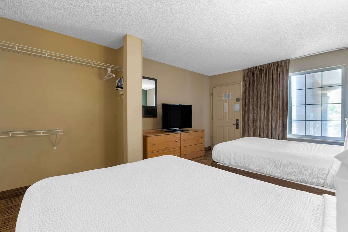 A hotel room with two beds, a wooden dresser, a television, and a window with curtains.