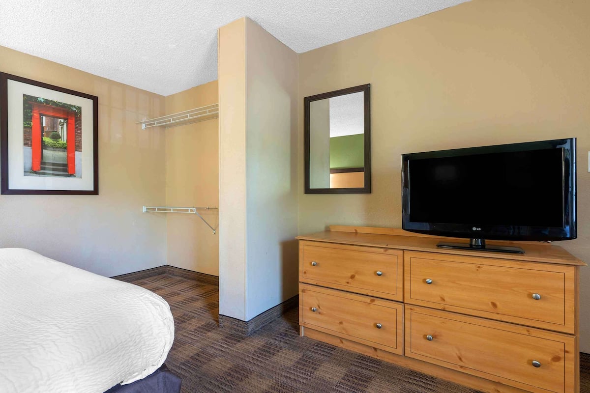 A hotel room with a bed, a wooden dresser, a flat-screen TV, a framed picture on the wall, and a mirror.