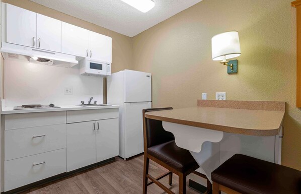 Cocina compacta con armarios blancos, microondas, refrigerador, una pequeña mesa con banco y una lámpara fijada a la pared.