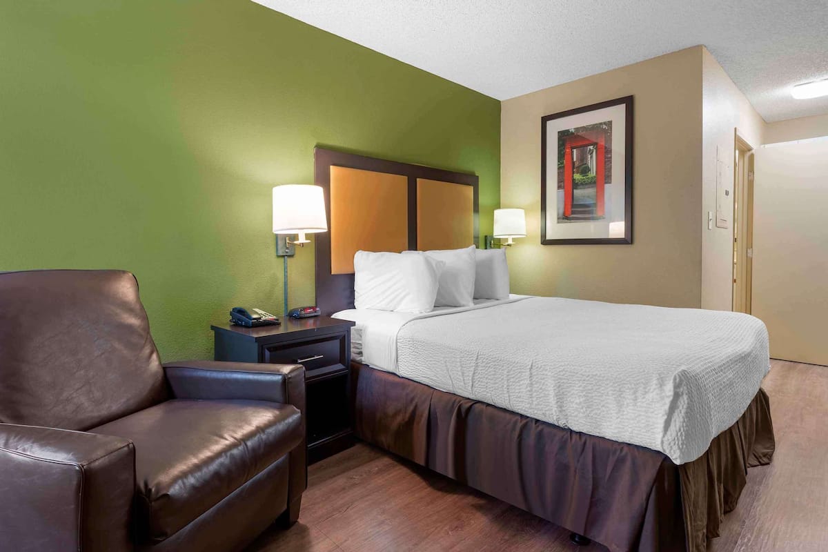 A hotel room with a bed, a brown leather chair, a nightstand with a lamp, and a framed picture on the wall.