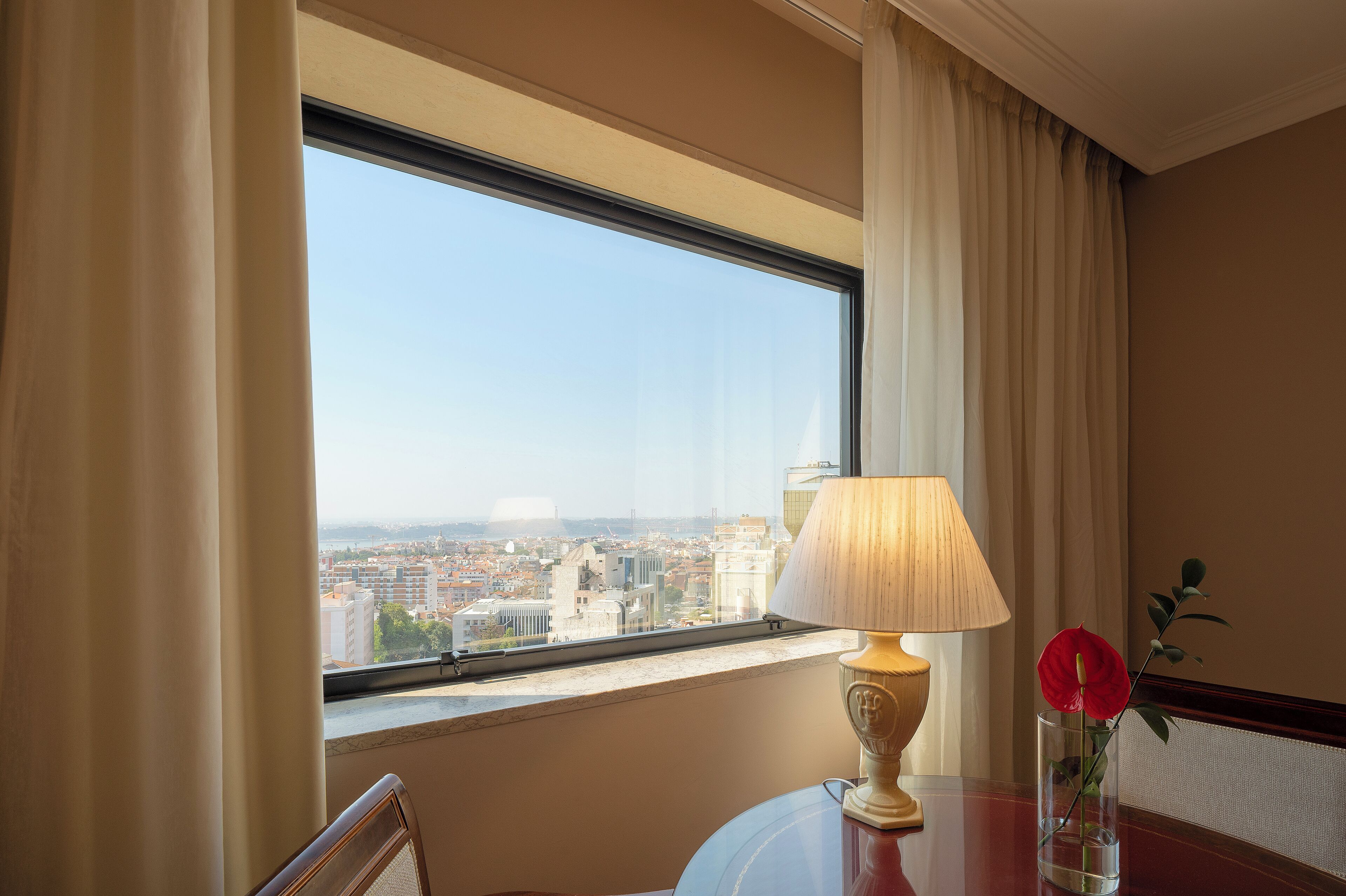Ein Hotelzimmer mit großem Fenster, einem Tisch mit Lampe und einer Vase mit einer Blume, und Blick auf die Stadtlandschaft.