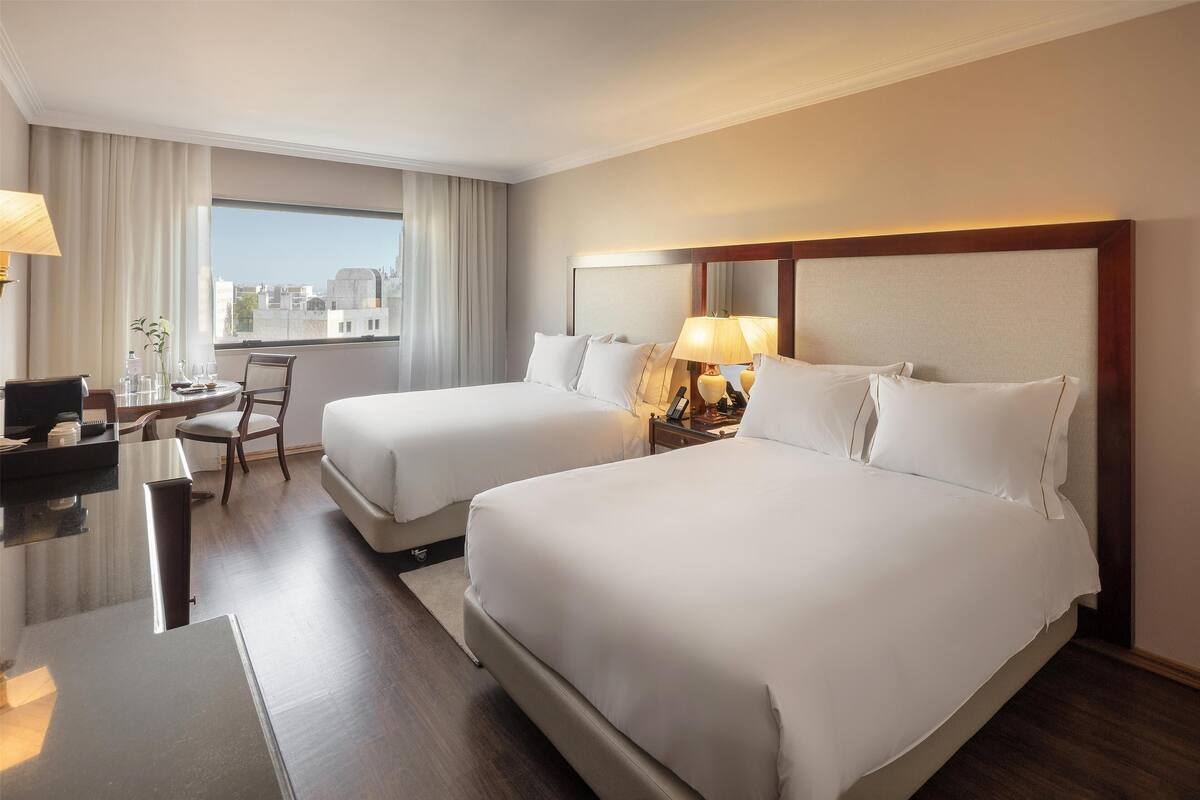 Habitación de hotel con dos camas, un escritorio, una silla y un amplio ventanal con vistas a la ciudad.