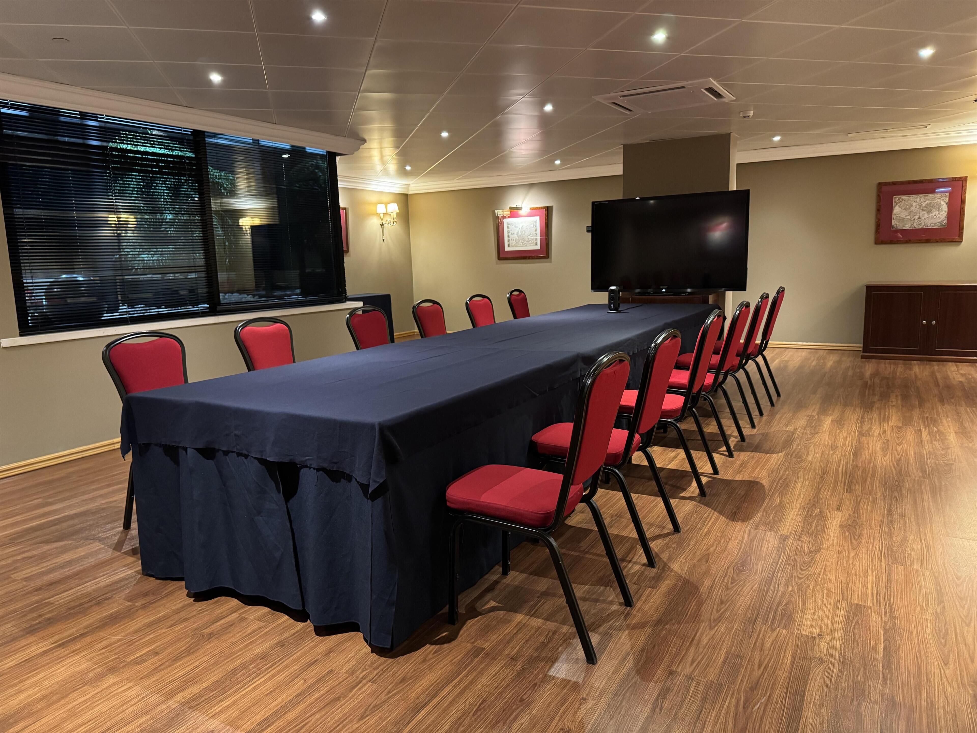 Una sala conferenze con un lungo tavolo, sedie rosse, un'ampia televisione a schermo piatto e una finestra con persiane.