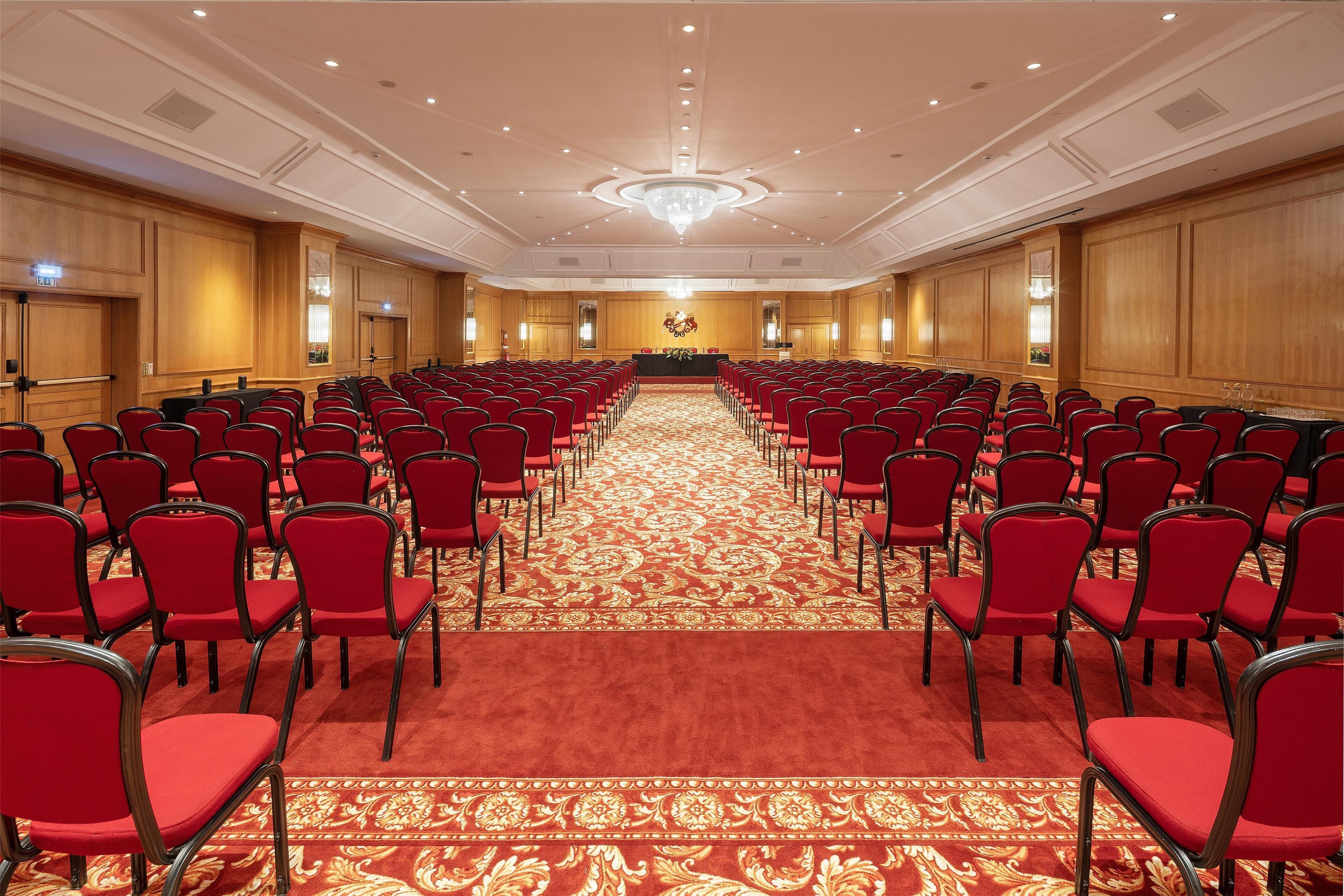 A conference room with rows of red chairs, a patterned carpet, and a chandelier.