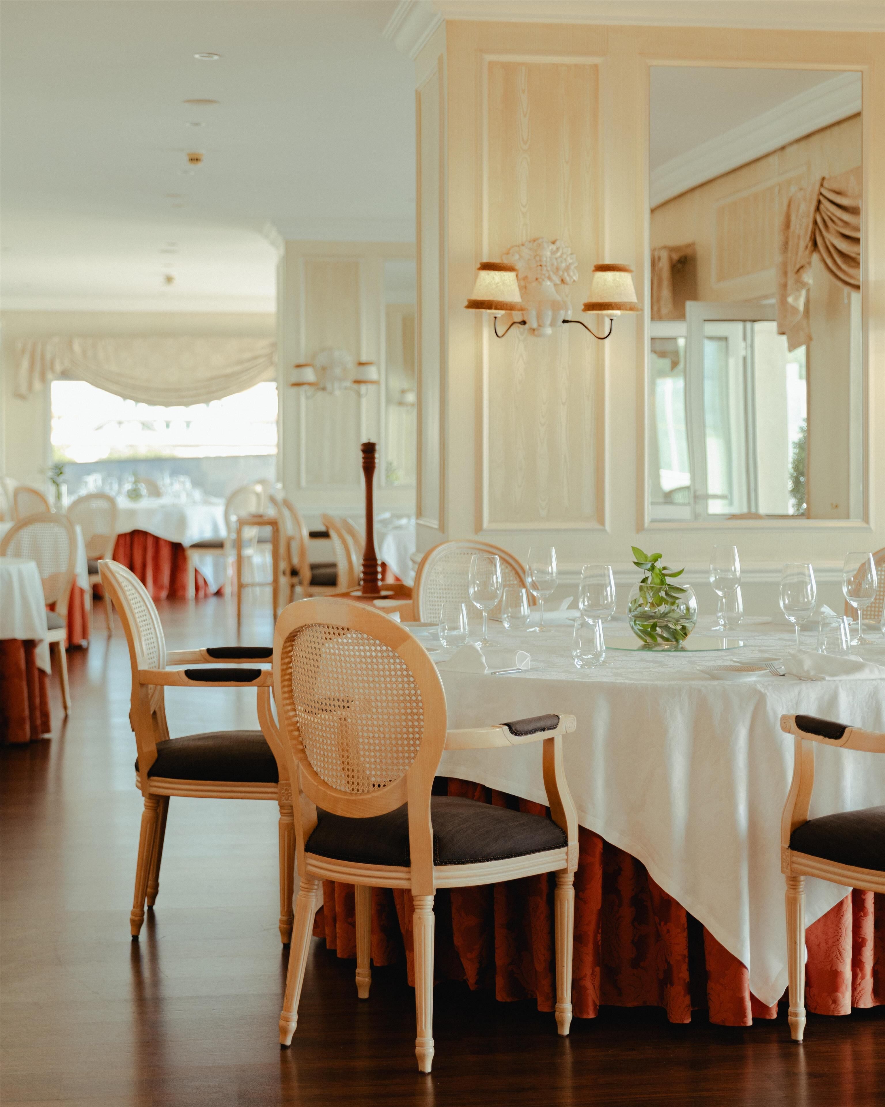 A dining area with round tables set for service, wooden chairs with cushioned seats, and a large mirror on the wall.