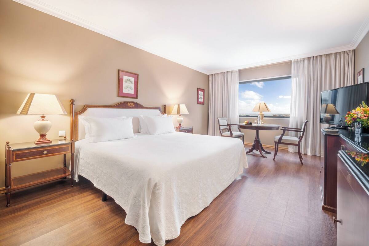 A hotel room with a large bed, two bedside tables with lamps, a desk with a chair, a TV, and a window with a view.