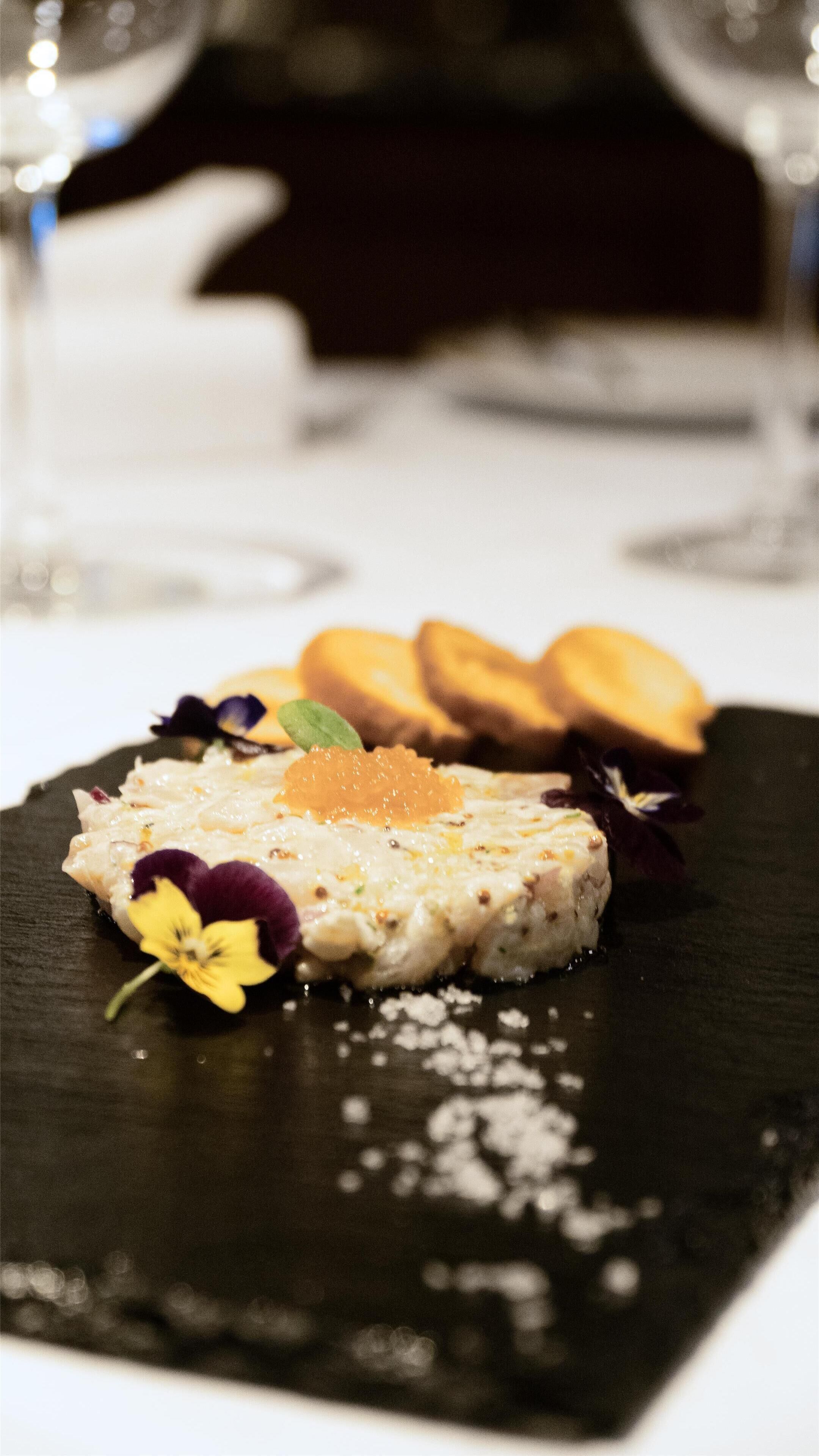A dessert plate with a creamy dish, a slice of fruit, and a flower on a dark slate.